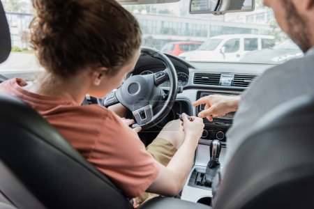 back view of father pointing with finger while showing teenage son how to start car 