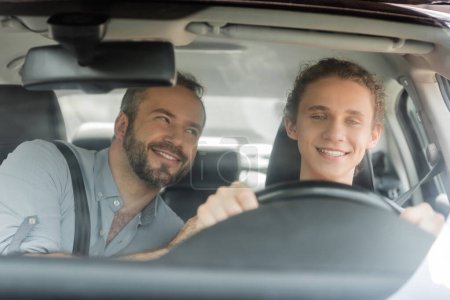 smiling teenager boy holding steering wheel while driving car next to dad 