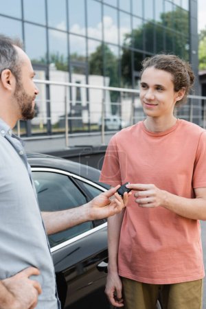 Glücklicher bärtiger Mann übergibt Autoschlüssel an fröhlichen Teenager-Sohn in der Nähe von Auto 