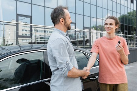 Foto de Hombre barbudo feliz estrechando la mano con alegre hijo adolescente sosteniendo la llave del coche cerca del automóvil - Imagen libre de derechos
