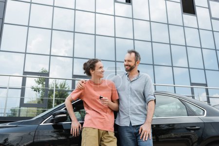 glücklicher bärtiger Mann legt Hand auf Schulter von fröhlichem Teenager-Sohn, der Autoschlüssel in der Nähe von Auto hält 