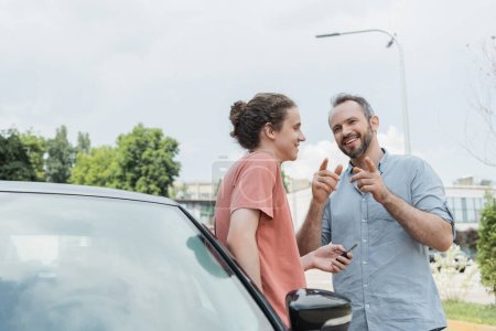 glücklicher bärtiger Vater zeigt mit den Fingern auf Teenager-Sohn, der neben Auto mit Schlüsseln steht 