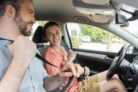 cheerful teenage boy and dad smiling while fastening safety belts in car 