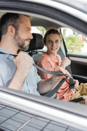 glücklicher Teenager und bärtiger Vater lächeln beim Anschnallen der Sicherheitsgurte im Auto 