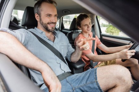 glücklicher bärtiger Vater zeigt mit dem Finger auf Teenager-Sohn, der Auto fährt 