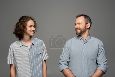 cheerful and bearded father looking at curly happy son isolated on grey 