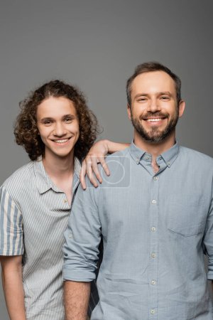 heureux père et fils en chemises regardant caméra et souriant isolé sur gris 