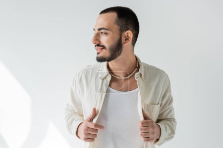Sonriente y barbudo hombre gay en ropa casual y collar de perlas tocando camisa y mirando hacia otro lado mientras posando sobre fondo gris con luz solar 
