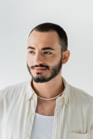Porträt eines jungen bärtigen homosexuellen Mannes in beigem Hemd, ärmellosem T-Shirt und Perlenkette, der isoliert auf grau steht und wegschaut 