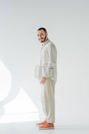 Longitud completa de hombre homosexual sonriente y barbudo en ropa beige con telas naturales mirando a la cámara mientras está de pie sobre fondo gris con luz solar 