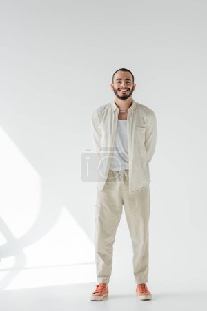 Full length of positive and short haired homosexual man in beige clothes and pearl necklace looking at camera while standing on grey background with sunlight