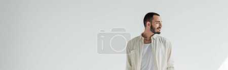 Smiling and young bearded gay man in casual clothes and trendy pearl necklace looking away while standing on grey background, banner 