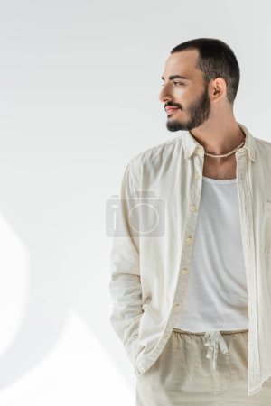 Homme homosexuel barbu et aux cheveux courts en chemise beige et collier de perles élégant détournant les yeux et tenant la main dans une poche de pantalon en tissus naturels sur fond gris avec lumière du soleil 