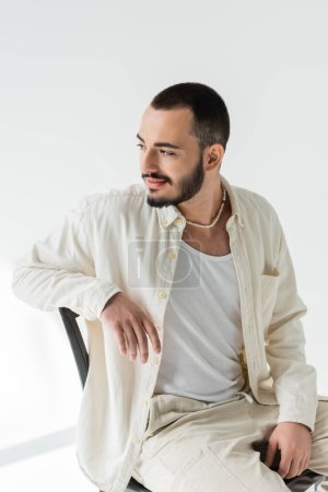 Young brunette homosexual man in beige clothes with natural fabrics and pearl necklace relaxing while sitting on chair on grey background with sunlight 