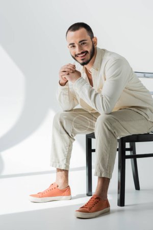 Full length of smiling short haired homosexual man in beige everyday clothes made of natural fabrics sitting on chair and looking at camera on grey background with light 