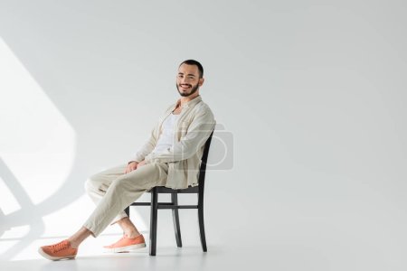 Morena positiva gay hombre en ropa casual hecha de telas naturales mirando a la cámara mientras está sentado en una silla cómoda sobre fondo gris con luz solar 