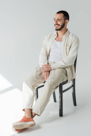 Carefree homosexual man in pearl necklace and casual clothes made of natural fabrics looking away while sitting on chair on grey background with sunlight 