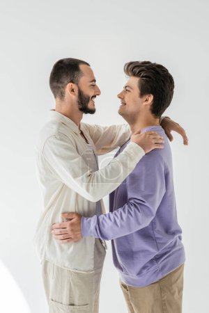 Foto de Vista lateral de pareja homosexual joven y alegre en ropa casual abrazándose y mirándose mientras están de pie sobre un fondo gris - Imagen libre de derechos