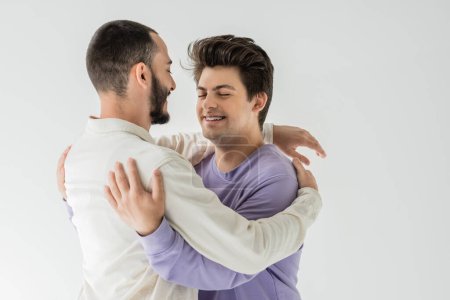 Morena llena de alegría pareja del mismo sexo en ropa casual abrazándose unos a otros y cerrando los ojos mientras está de pie aislado en gris 