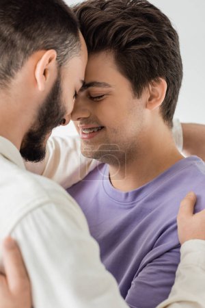 Portrait of positive brunette gay man in braces hugging bearded partner in shirt while closing eyes and standing nose to nose isolated on grey 