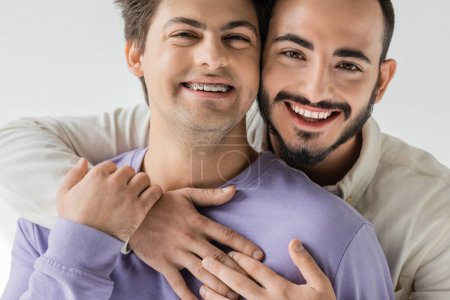 Foto de Retrato de pareja joven y alegre del mismo sexo en ropa casual abrazando y tocando las manos mientras mira la cámara aislada en gris - Imagen libre de derechos