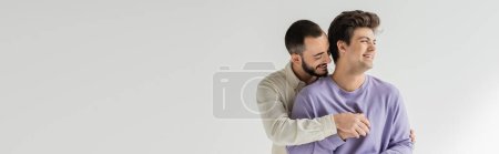 Foto de Joven hombre homosexual barbudo abrazando despreocupado novio morena con frenillos y sudadera mirando hacia otro lado mientras está de pie aislado en gris, pancarta - Imagen libre de derechos