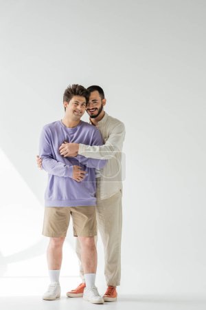Téléchargez les photos : Pleine longueur de couple positif de même sexe dans des vêtements décontractés tenant la main et regardant la caméra tout en se tenant sur fond gris avec la lumière du soleil - en image libre de droit
