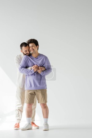 Téléchargez les photos : Homme homosexuel souriant et barbu en vêtements décontractés tenant la main d'un jeune petit ami les yeux fermés tout en se tenant sur fond gris avec la lumière du soleil - en image libre de droit
