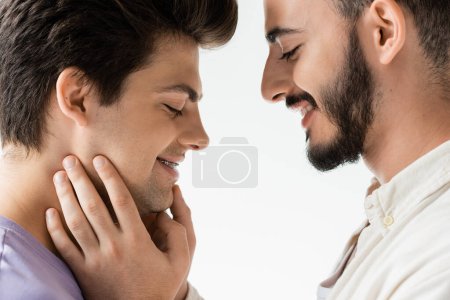 Foto de Vista lateral del hombre gay barbudo positivo en camisa casual tocando la cara de la joven pareja con tirantes en los dientes mientras están de pie juntos aislados en gris - Imagen libre de derechos