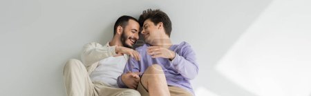 Foto de Riendo pareja homosexual con los ojos cerrados tomados de la mano y conversando mientras están sentados juntos sobre un fondo gris con luz solar, pancarta - Imagen libre de derechos