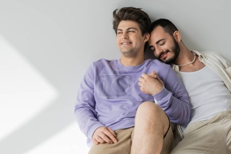 Smiling young gay man in casual clothes holding hand of bearded boyfriend with closed eyes while sitting together on grey background with sunlight 
