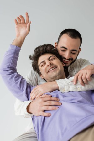 Positiver und bärtiger homosexueller Mann in lässiger Kleidung umarmt brünetten Partner mit geschlossenen Augen und Zahnspange, während er isoliert auf grau sitzt 