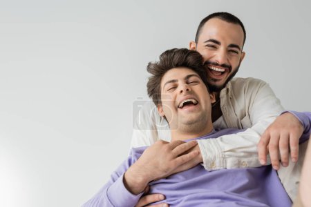 Bearded homosexual man in shirt embracing and touching hand of laughing boyfriend in braces and purple sweatshirt while sitting isolated on grey 