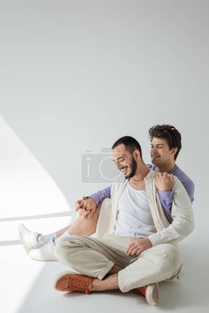 Young gay man smiling while holding hand and hugging bearded boyfriend in casual shirt while sitting on grey background with sunlight 