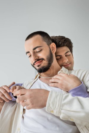 Portrait of smiling and bearded homosexual man in casual clothes and pearl necklace touching hand of brunette boyfriend closing eyes isolated on grey 