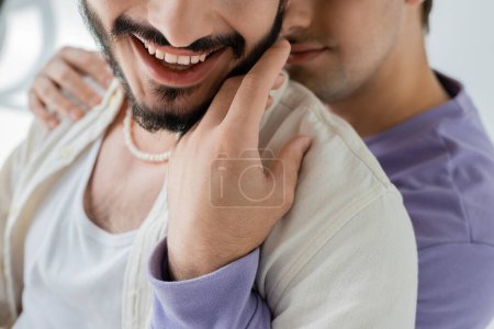 Cropped view of blurred homosexual man in casual clothes hugging and touching beard of carefree partner in casual clothes on grey background