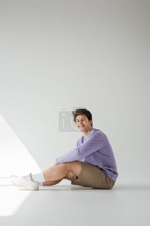 Full length of smiling homosexual man in braces, beige shorts and purple sweatshirt looking at camera while sitting on grey background with sunlight 