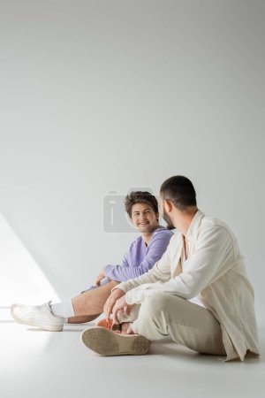 Téléchargez les photos : Pleine longueur de sourire homme homosexuel en sweat-shirt violet regardant copain barbu tout en étant assis ensemble sur fond gris avec la lumière du soleil - en image libre de droit