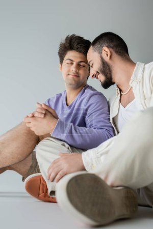 Young and positive same sex couple with closed eyes in casual clothes holding hands while sitting and resting together on grey background