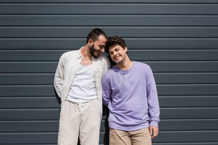 Lächelnder und bärtiger schwuler Mann in lässiger Kleidung, der neben einem jungen Freund in Hosenträgern steht und tagsüber in Kamera und Wand eines grauen Gebäudes im Freien blickt 
