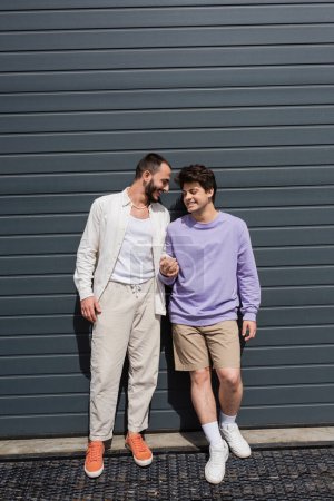 Foto de Longitud completa de pareja positiva y joven del mismo sexo en ropa casual tomados de la mano mientras están de pie juntos cerca del edificio gris en la calle urbana - Imagen libre de derechos
