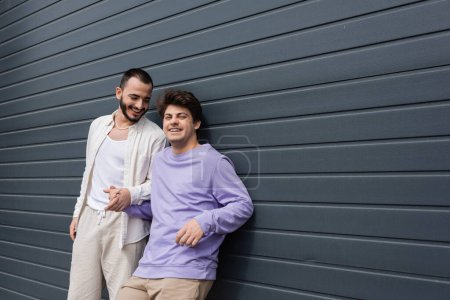 Cheerful and young gay man in sweatshirt and braces holding hand of bearded boyfriend while standing together near wall of building on urban street 