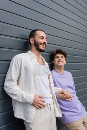 Homosexual bearded man smiling and looking away while holding hand of young boyfriend in braces near wall of building outdoors at daytime