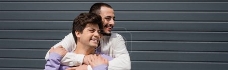 Young bearded man hugging smiling gay partner in casual clothes and looking away while standing near building outdoors, banner 