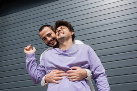 Low angle view of positive and bearded gay man hugging brunette boyfriend in sweatshirt and braces while looking at camera near building on urban street 