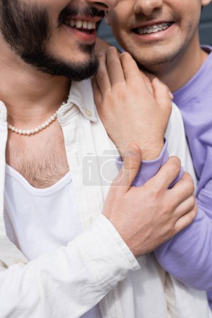 Cropped view of carefree homosexual man in purple sweatshirt with braces hugging and touching shoulder of bearded partner outdoors at daytime