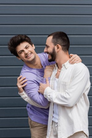 Portrait of cheerful homosexual man in purple sweatshirt and braces hugging young bearded boyfriend and looking at camera while standing near building outdoors 
