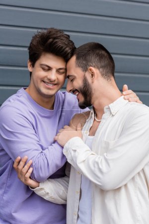Portrait of positive homosexual man in casual clothes and braces hugging bearded boyfriend in shirt and pearl necklace with closed eyes while standing near building outdoors 