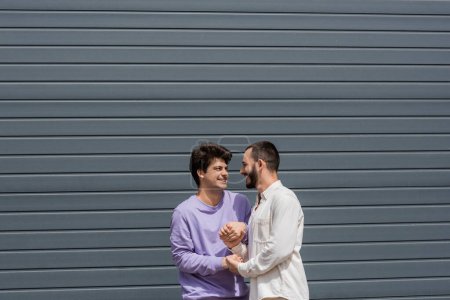 Foto de Pareja homosexual positiva y joven tomados de la mano mientras hablan y se miran el uno al otro cerca del edificio en la calle urbana durante el día - Imagen libre de derechos