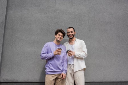 Smiling and young same sex couple in casual clothes holding coffee to go and looking at camera while standing near building outdoors 